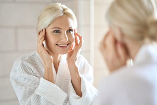 mid age adult 50 years old blonde woman standing in bathroom after shower touching face 1 1